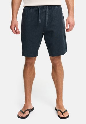 Homme portant un short bleu marine à cordon de serrage et des tongs noires, debout avec les bras détendus le long du corps sur fond blanc.