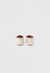 Ζευγάρι μπεζ δερμάτινα μοκασίνια με καφέ δερμάτινες λεπτομέρειες και φιόγκο, εμφανιζόμενα από πίσω σε απλό λευκό φόντο.