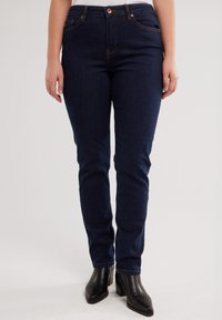 Naine, kes kannab tumedat sinist kitsast teksapükste ja musti pahkluu-saapad, seisab neutraalse tausta ees.