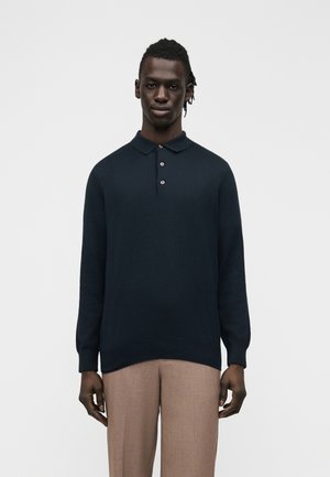 Férfi, sötétkék hosszú ujjú piké pulóvert és világosbarna nadrágot visel, egy egyszerű háttér előtt állva, előre nézve.