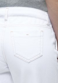 Άτομο που φοράει λευκό τζιν από denim και γκρι τοπ, δείχνοντας την πίσω τσέπη και την περιοχή της μέσης του τζιν.