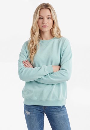 Młoda kobieta z długimi blond włosami, ubrana w jasnoniebieską bluzę i podarte niebieskie dżinsy, stoi z założonymi ramionami na białym tle.