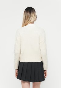 Jersey blanco de punto trenzado con un dobladillo acanalado, combinado con una falda negra con pliegues. El jersey presenta mangas texturizadas y un diseño de cuello clásico.