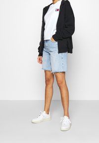 Svart dragkedjehuva över en vit t-shirt, matchad med ljusblå denimshorts med fransar och vita sneakers. Enkelt, avslappnat outfit.