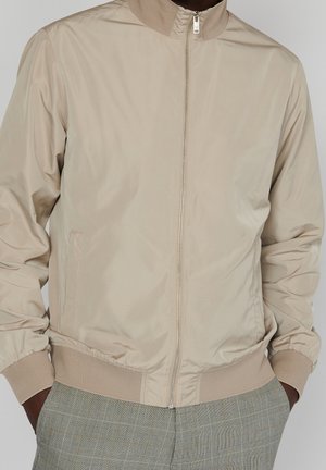 Mann trägt beige Zip-up-Bomberjacke mit gerippten Bündchen und Kragen, kombiniert mit grauen karierten Hosen, Hände in den Taschen.