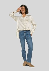 Silk-like cream blouse, blue denim jeans, beige ankle boots with accents. Model poses with hand on head against a gray background.