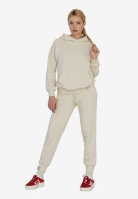 Beżowy dres składający się z bluzy z kapturem i spodni dresowych wykonany z miękkiego materiału; posiada kieszeń kangurką i elastyczne mankiety; zestawiony z czerwonymi trampkami.