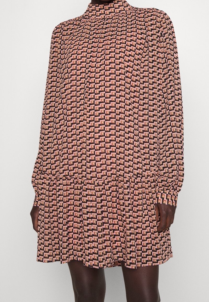 Robe à manches longues et col haut avec motif géométrique rose et noir, coupe ample et ourlet à volants, portée par une personne à la peau foncée.
