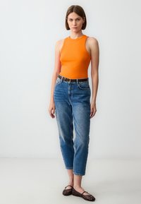 Top arancione, jeans blu a vita alta con dettagli sbiaditi, cintura marrone e ballerine intrecciate marroni su uno sfondo semplice.