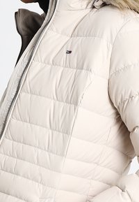 Ljusbeige pufferjacka med horisontell quiltad design, dragkedjestängning och en huva med pälsfoder. Har en liten logotyp på bröstet.