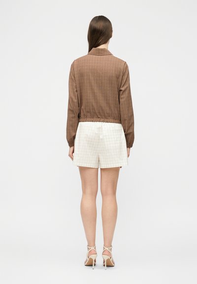 Γυναίκα όρθια, με όψη από πίσω, φοράει cropped καφέ μακρυμάνικο μπουφάν, λευκά σορτς και μπεζ πέδιλα με ψηλό τακούνι σε απλό φόντο.