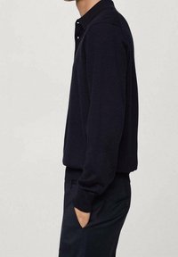 Osoba ubrana w ciemnoniebieską bluzę z długim rękawem i pasujące spodnie, stojąca na jednolitym tle z jedną ręką w kieszeni.