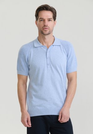 Hombre con cabello corto y castaño, vistiendo una camiseta polo azul claro de manga corta y pantalones oscuros, de pie frente a un fondo neutro.