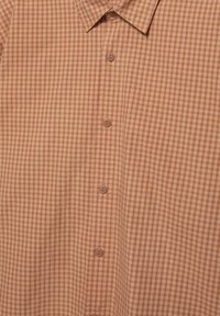 Ruutupaita maanläheisissä ruskean ja oranssin sävyissä, jossa nappikiinnitys ja vaaleanruskeat napit sekä klassinen kaulus.