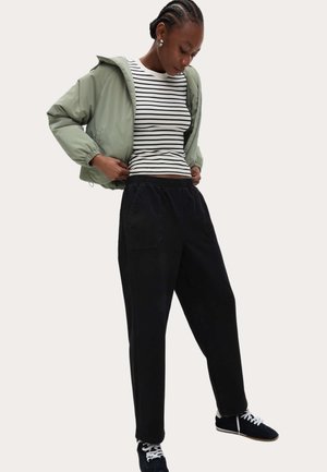 Jonge persoon stelt lichtgroene capuchonjas bij, draagt zwarte losse broek, zwart-witte sneakers en een wit shirt met zwarte horizontale strepen.