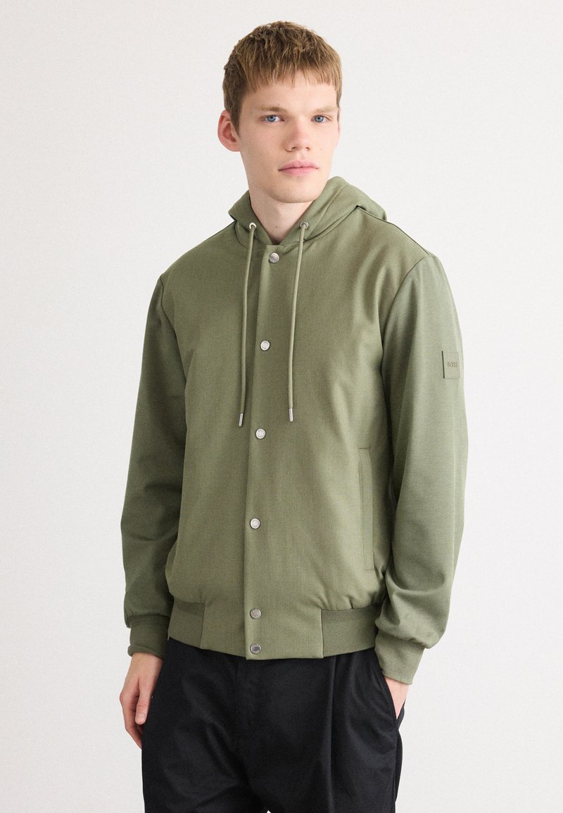 Chaqueta con capucha de color verde oliva, hecha de un tejido suave, que presenta un cierre frontal de botones, capucha ajustable y bolsillos laterales. Diseño sencillo con detalle de logo.