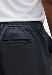 Κοντινή λήψη της ζώνης μέσης ενός ατόμου που φοράει μαύρο παντελόνι με ελαστική μέση, τσέπη με σχισμή στο πίσω μέρος και μια μικρή υφασμάτ ινη ετικέτα κοντά στην άκρη της τσέπης.