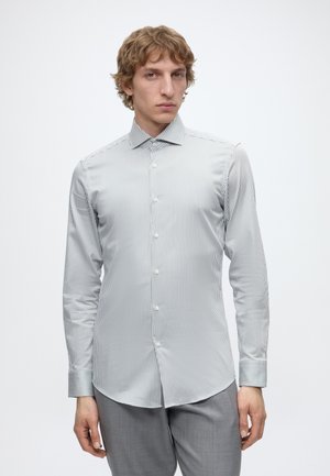 Νέος άνδρας που φοράει άσπρο πουκάμισο slim-fit με λεπτές κάθετες ρίγες και γκρι παντελόνι, στέκεται μπροστά σε απλό λευκό φόντο.