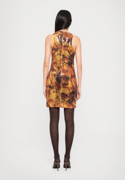 Woman wearing a sleeveless, sequined orange and brown patterned mini dress, black tights, and brown high-heeled shoes, standing with her back facing forward.