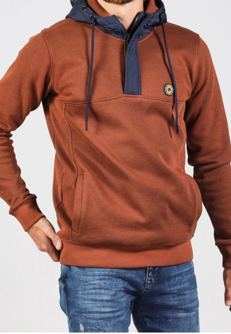 Sweat à capuche marron avec capuche bleu marine et cordons, porté avec un jean bleu, montrant le haut du corps et la main dans la poche.