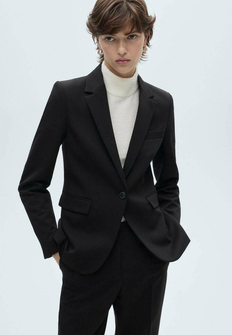 Černý přizpůsobený blazer s jedním knoflíkem, bočními kapsami a saténovou Texturou. Nosený přes světlý rolák. Minimalistický design, bez vzorů.
