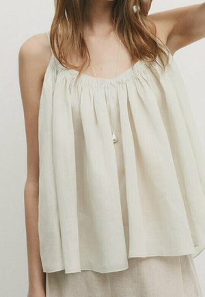 Haut ample sans manches crème avec encolure froncée porté par une personne aux cheveux longs, associé à un bas de couleur claire et un collier pendentif.