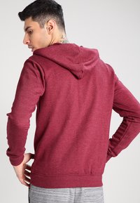 Burgunderfarget hettegenser laget av mykt stoff, med snor i hetten, lange ermer og ribbestrikket kant. Glatt tekstur med minimalt synlig søm.