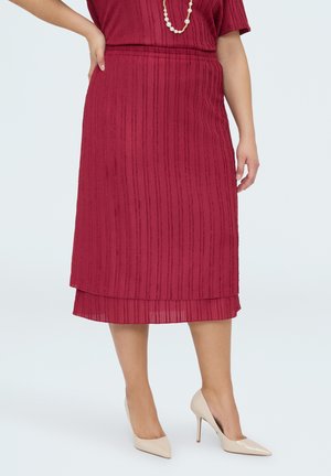 Kvinde iført en rød tekstureret midinederdel, en matchende top, perlekæde og beige højhælede sko, stående med den ene hånd på hoften.