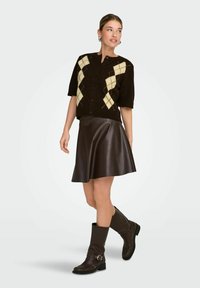 Cardigan marron à manches courtes avec un motif argyle crème, associé à une jupe en cuir marron de style trapèze et des bottes marron montant jusqu'au genou avec une boucle.