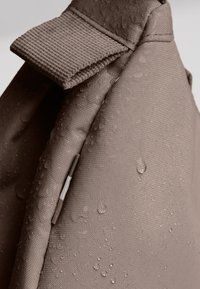 Primer plano de la esquina de una mochila de tela beige con gotas de agua en su superficie y costuras reforzadas en el lazo de la correa.