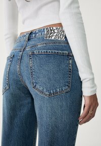 Los vaqueros de tiro alto en denim azul presentan un tejido texturizado, diseño de cinco bolsillos y una etiqueta en contraste en blanco y negro en la cintura trasera.
