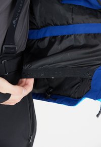 Interno di giacca a vento nera con accenti in tessuto blu e nero. Presenta cinghie elastiche e una texture liscia; la fodera interna mostra i dettagli della cucitura.