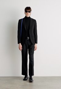 Zwart op maat gemaakt pak met een hoge zwarte coltrui, slim-fit broek en zwarte leren schoenen. Blauwe schoudertas en zonnebril.
