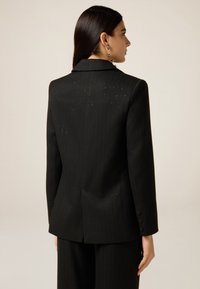 Czarna marynarka w prążki o usztywnionej konstrukcji, z kołnierzem z wycięciem, mankietami na trzy guziki i rozcięciem na środku z tyłu.