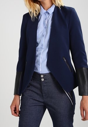 Femme portant une veste bleu marine ajustée avec des poignets en cuir noir, une chemise bleue claire boutonnée, et un pantalon bleu foncé texturé avec des doubles boutons argentés.