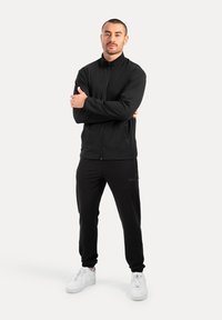 Schwarze Zip-Jacke mit hohem Kragen, kombiniert mit schwarzen Hosen. Sie hat eine glatte Textur und weiße Sneakers. Minimalistisches Design.