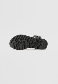 Schwarze Sandalensohle mit einer strukturierten Gummiaußensohle, die Zickzack-Profilmuster aufweist. Enthält eine metallische Schnallenakzent und ein Obermaterial aus Leder.