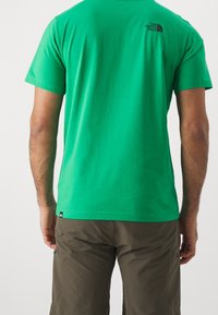 Vihreä puuvillapaita lyhyillä hihoilla, jossa on musta logo yläselässä, yhdistettynä khaki-shorteihin. Sileä kangas.