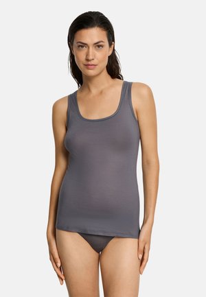 Vrouw met donker haar, gekleed in een nauwsluitend grijs hemdje en bijpassend ondergoed, staande tegen een effen witte achtergrond.