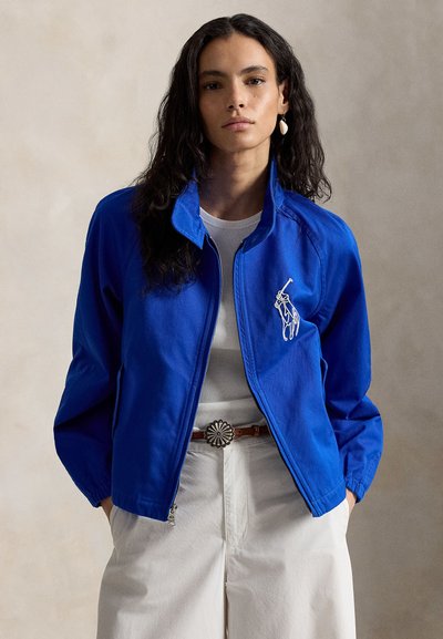 Vrouw met lang donker haar, gekleed in een felblauwe jas met ritssluiting over een wit shirt, witte broek en een bruine riem met een zilveren gesp.