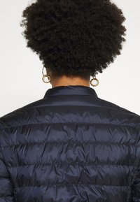 Person with curly black hair wearing gold hoop earrings and a dark navy quilted jacket, viewed from behind against a white background.