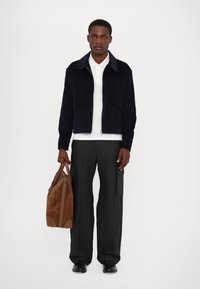 Navy corduroy jacket with two front pockets, white polo shirt, gray wide-leg trousers, and a brown suede duffel bag.