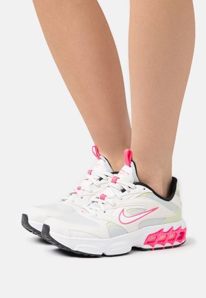 Pieds chaussés de baskets blanches avec des accents roses, des panneaux en mesh et des semelles rembourrées sur un fond uni.