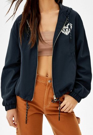 Femme portant un sweat à capuche noir zippé avec cordons de serrage, un crop top beige et un pantalon taille haute couleur rouille avec un bouton et des poches.