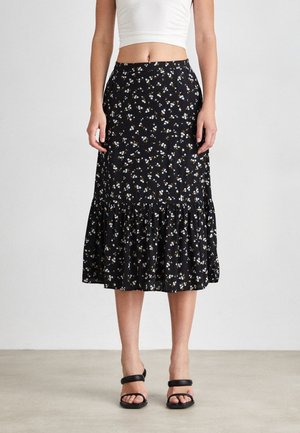 Frau trägt einen schwarzen Midirock mit weißem und gelbem Blumenmuster, kombiniert mit einem weißen Crop-Top und schwarzen Sandalen mit Absatz.