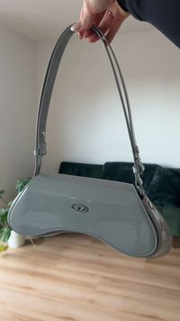 Grå, glansig handväska med en böjd form och justerbar rem. Har en central logo och en slät yta, placerad mot en minimalistisk bakgrund.