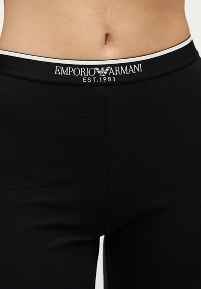 Gros plan du bas du torse d'une personne portant un sous-vêtement noir Emporio Armani avec un logo blanc et un texte sur la ceinture.