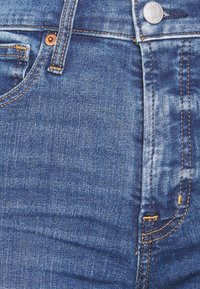 Jeans de mezclilla azul con cremallera, botón de cobre y costuras amarillas en contraste. El material tiene una textura suave con un ligero desgaste.