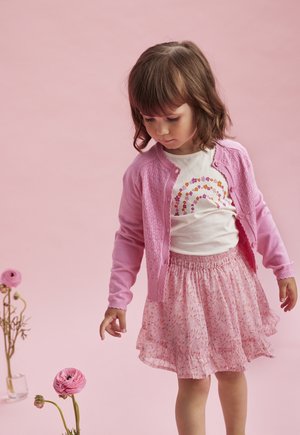 Lyserød cardigan med tekstureret design, en hvid skjorte med et farverigt regnbueprint og en luftig lyserød blomsterskørt med volanter.