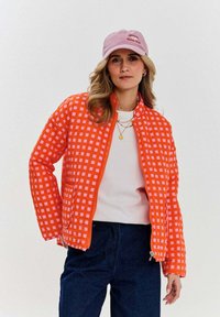 Prešívaná bunda v jasnej oranžovej farbe s bielym a ružovým mriežkovaným vzorom. Obsahuje zips a dve predné vrecká. Nosí sa s ľahkým topom a modrými džínsami.